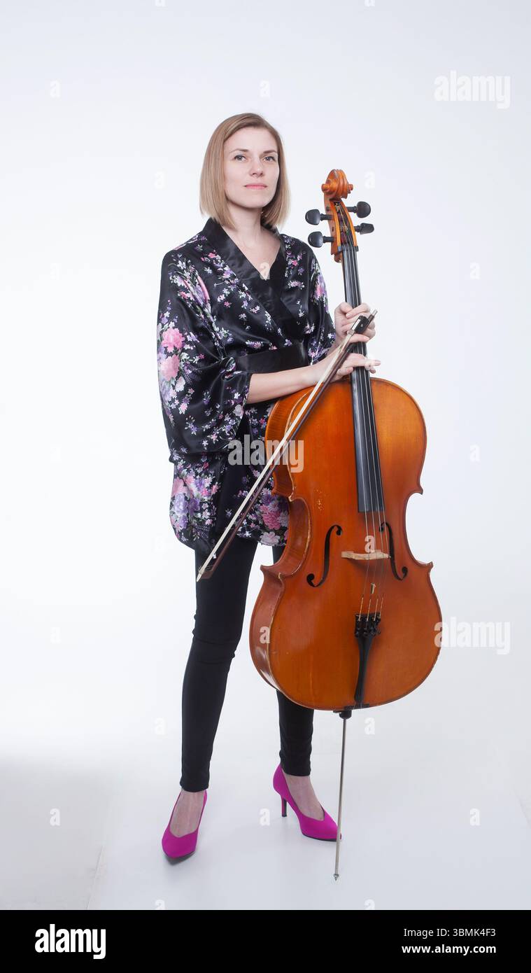 Donna caucasica adulta in uno studio con violoncello e arco. Indossa un abito nero in stile kimono floreale, leggings neri e un alto rosa brillante Foto Stock