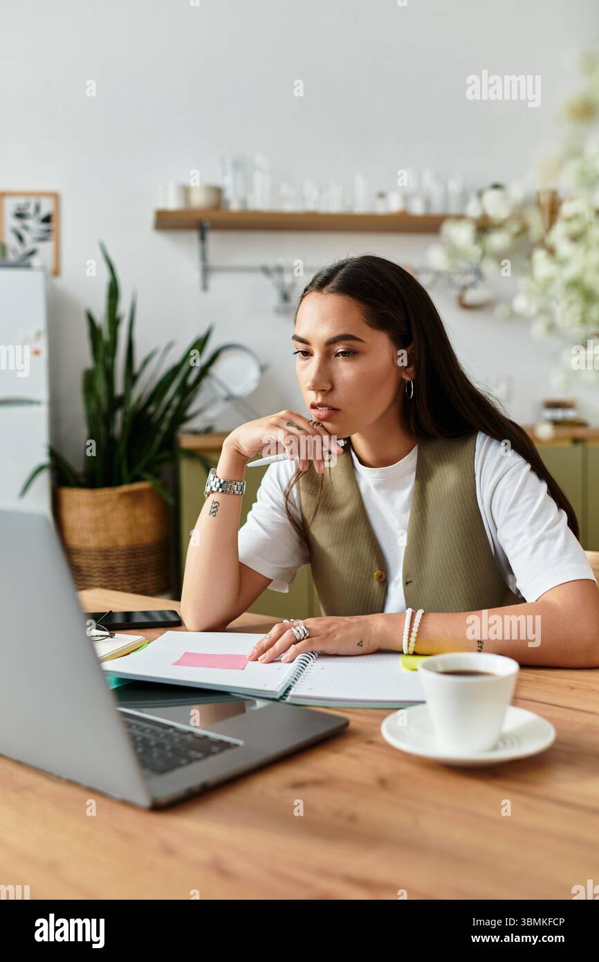 Una bella giovane donna siede comodamente alla sua scrivania, si concentra sul suo laptop e si gusta un caffè. Foto Stock