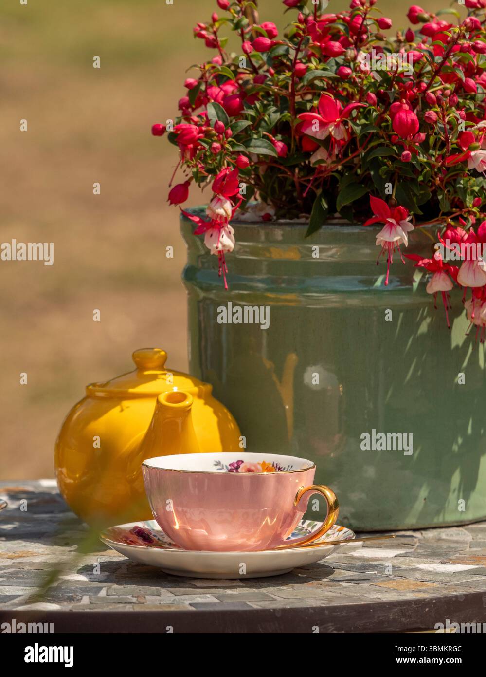 Un tè vintage rosa e oro con una teiera gialla brillante su un tavolo a mosaico accanto a un vaso di fucsia fiorita, crogiolandosi al sole estivo. Foto Stock