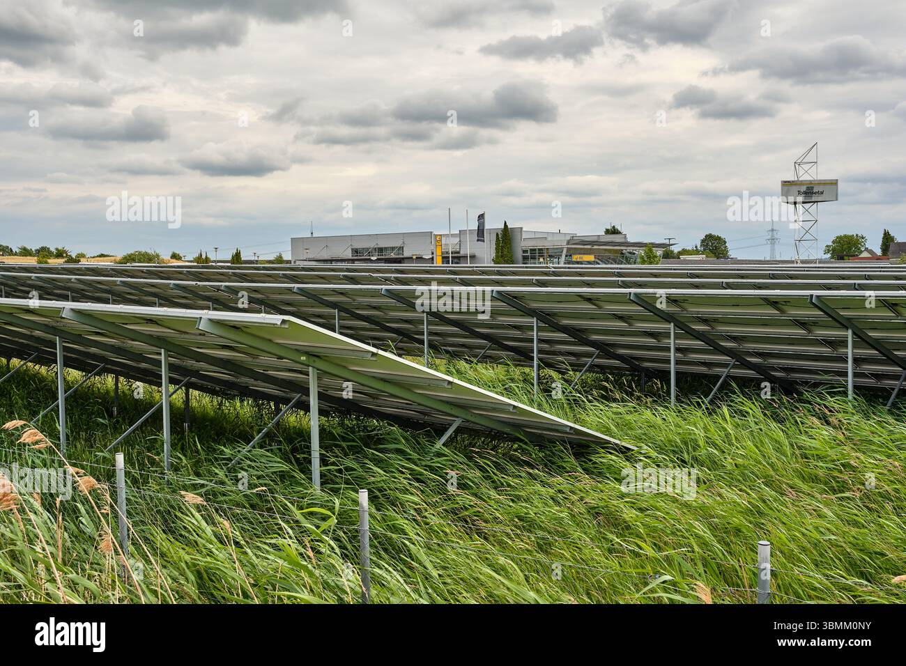 25 giugno 2024 - Altentreptow-Germania: File di pannelli solari su un campo verde vicino a un sito industriale sotto un cielo nuvoloso nel Meclemburgo, evidenziando re Foto Stock
