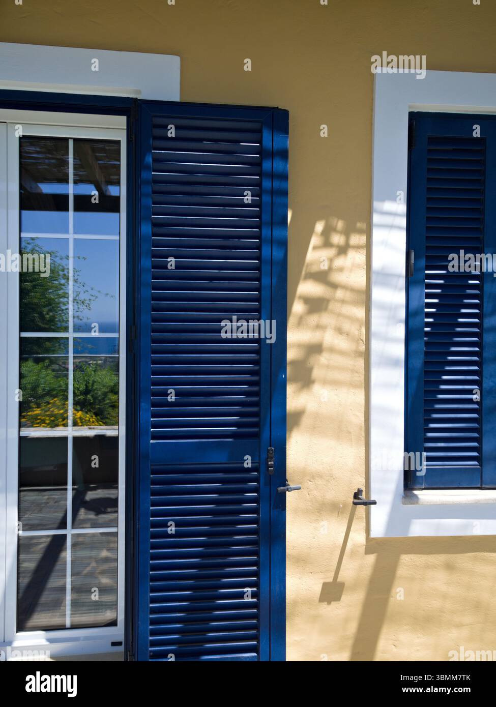 Facciata gialla della casa con persiane blu aperte sulla finestra e porta del balcone sotto la luce del sole. Foto Stock