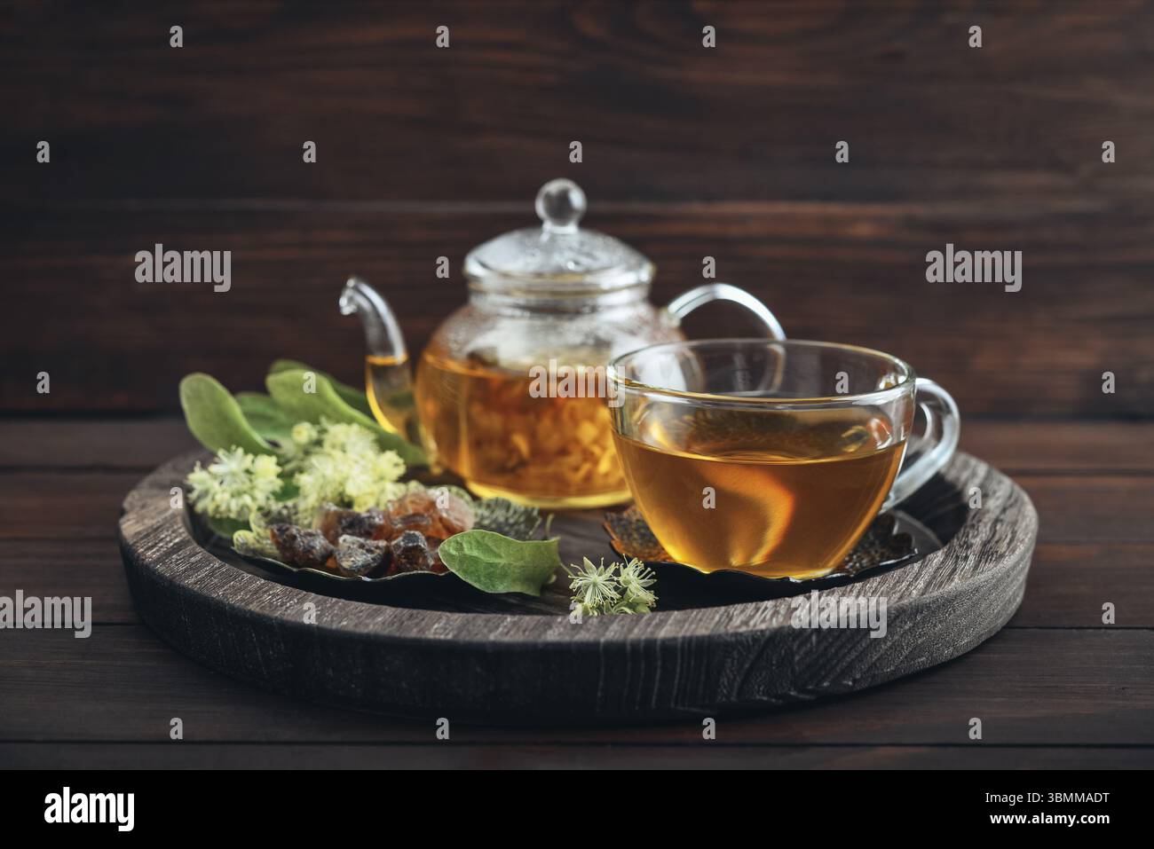 Tazza di tè al tiglio con teiera di vetro, zucchero e fiori di tiglio su vassoio rotondo su sfondo in legno Foto Stock