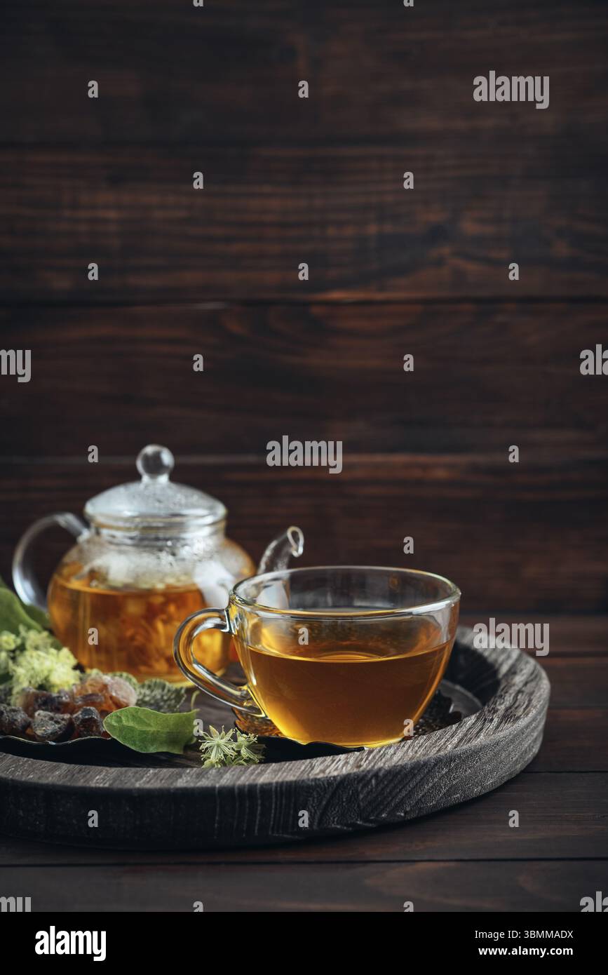 Tazza di tè al tiglio con teiera di vetro, zucchero e fiori di tiglio su vassoio rotondo su sfondo in legno Foto Stock