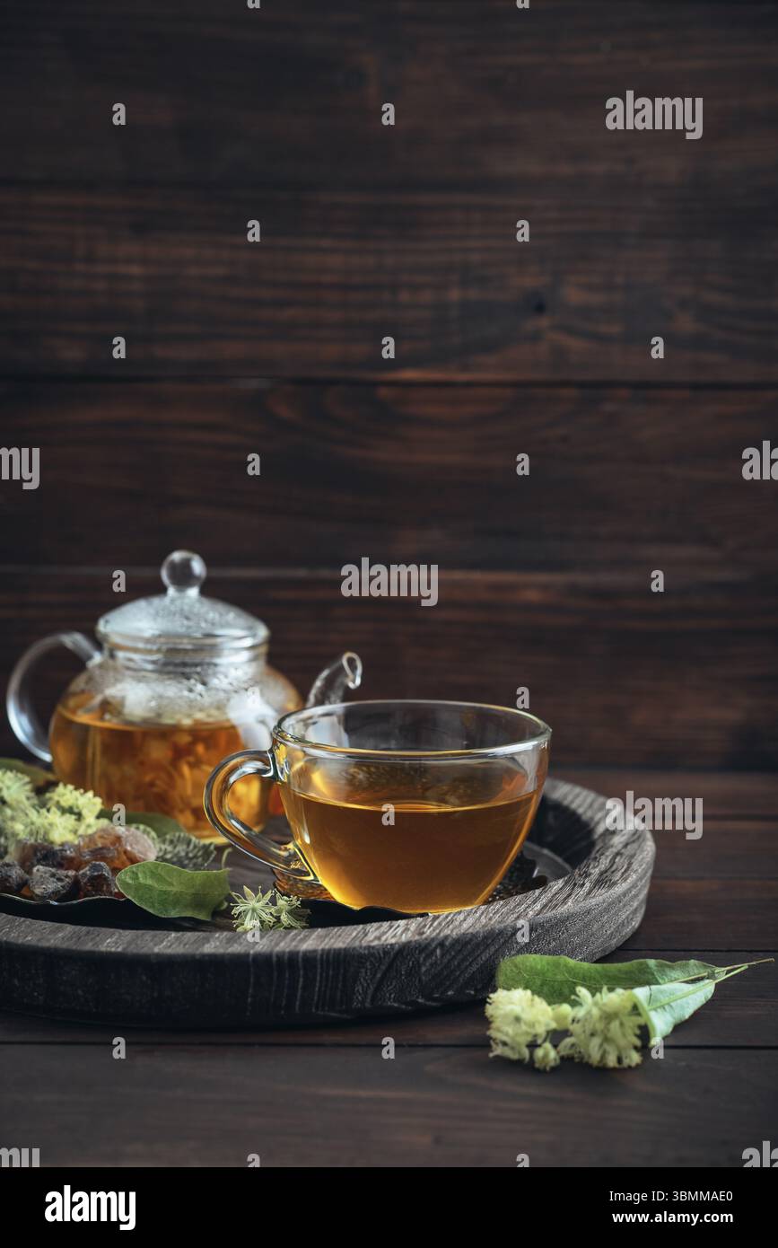 Tazza di tè al tiglio con teiera di vetro, zucchero e fiori di tiglio su vassoio rotondo su sfondo in legno Foto Stock