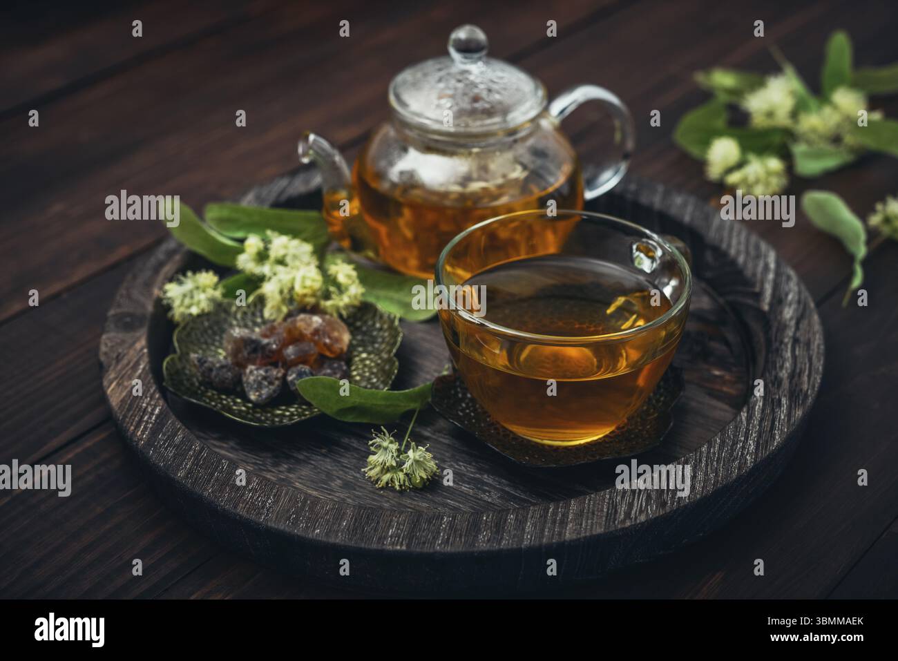 Tazza di tè al tiglio con teiera di vetro, zucchero e fiori di tiglio su vassoio rotondo su sfondo in legno Foto Stock