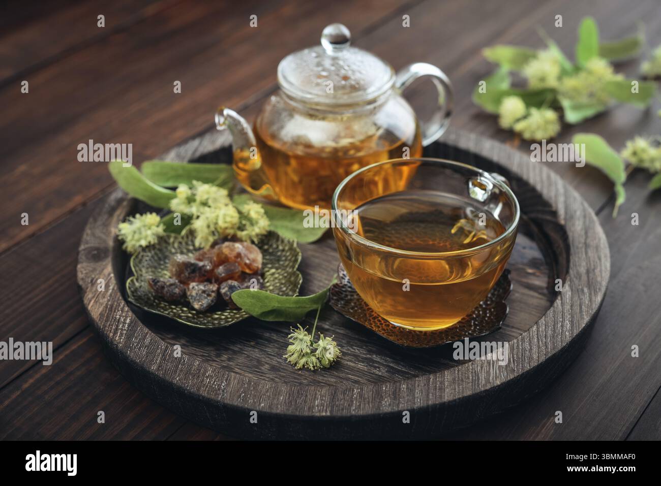 Tazza di tè al tiglio con teiera di vetro, zucchero e fiori di tiglio su vassoio rotondo su sfondo in legno Foto Stock