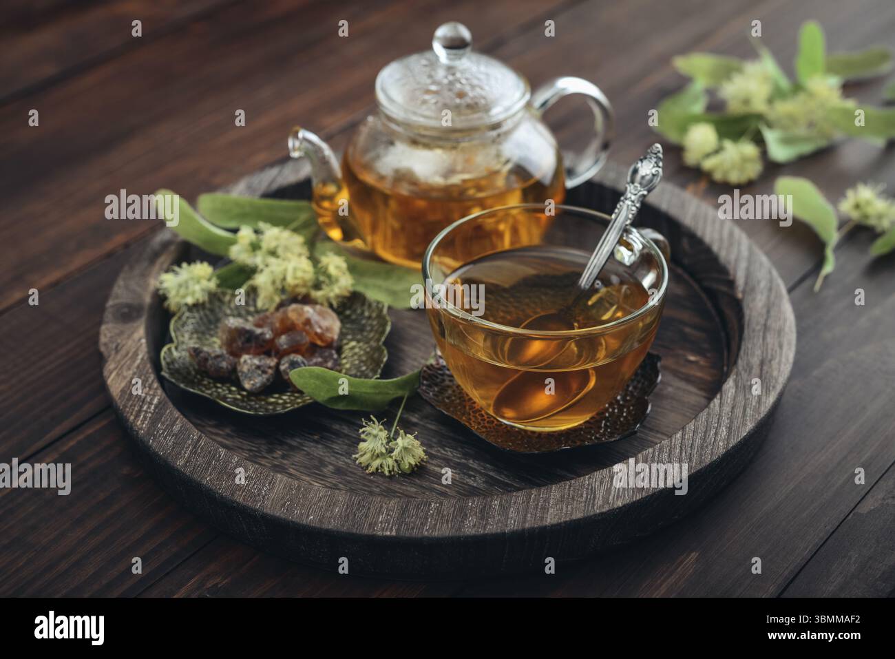 Tazza di tè al tiglio con teiera di vetro, zucchero e fiori di tiglio su vassoio rotondo su sfondo in legno Foto Stock