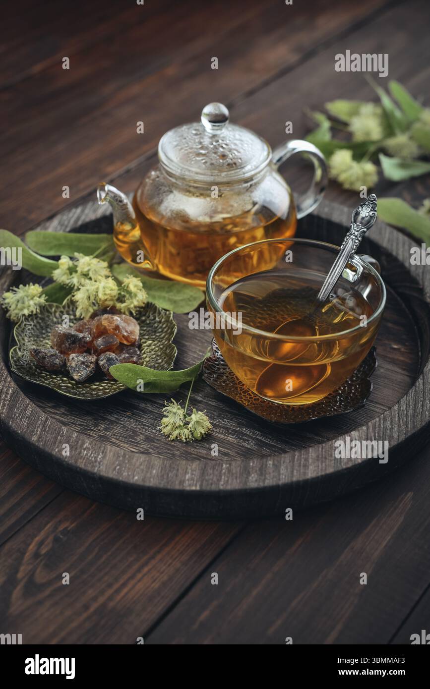 Tazza di tè al tiglio con teiera di vetro, zucchero e fiori di tiglio su vassoio rotondo su sfondo in legno Foto Stock