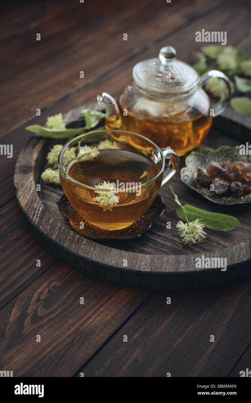 Tazza di tè al tiglio con teiera di vetro, zucchero e fiori di tiglio su vassoio rotondo su sfondo in legno Foto Stock