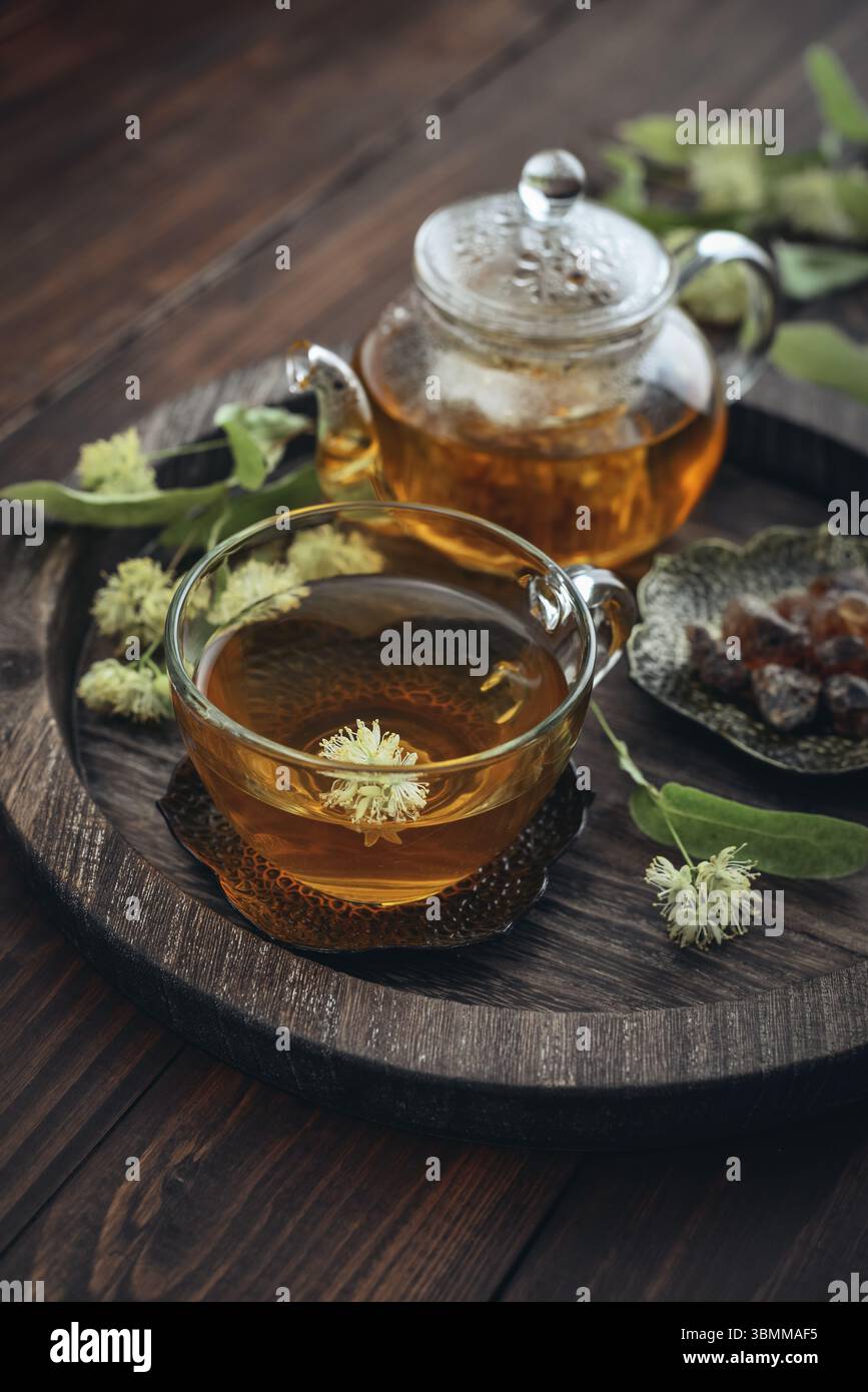 Tazza di tè al tiglio con teiera di vetro, zucchero e fiori di tiglio su vassoio rotondo su sfondo in legno Foto Stock