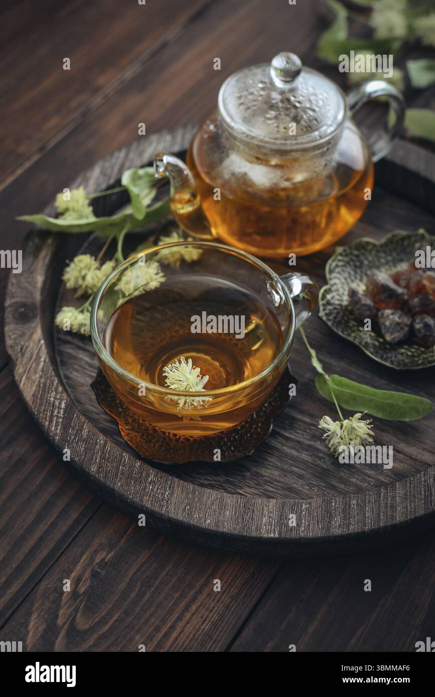 Tazza di tè al tiglio con teiera di vetro, zucchero e fiori di tiglio su vassoio rotondo su sfondo in legno Foto Stock