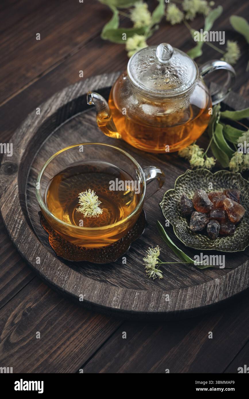 Tazza di tè al tiglio con teiera di vetro, zucchero e fiori di tiglio su vassoio rotondo su sfondo in legno Foto Stock