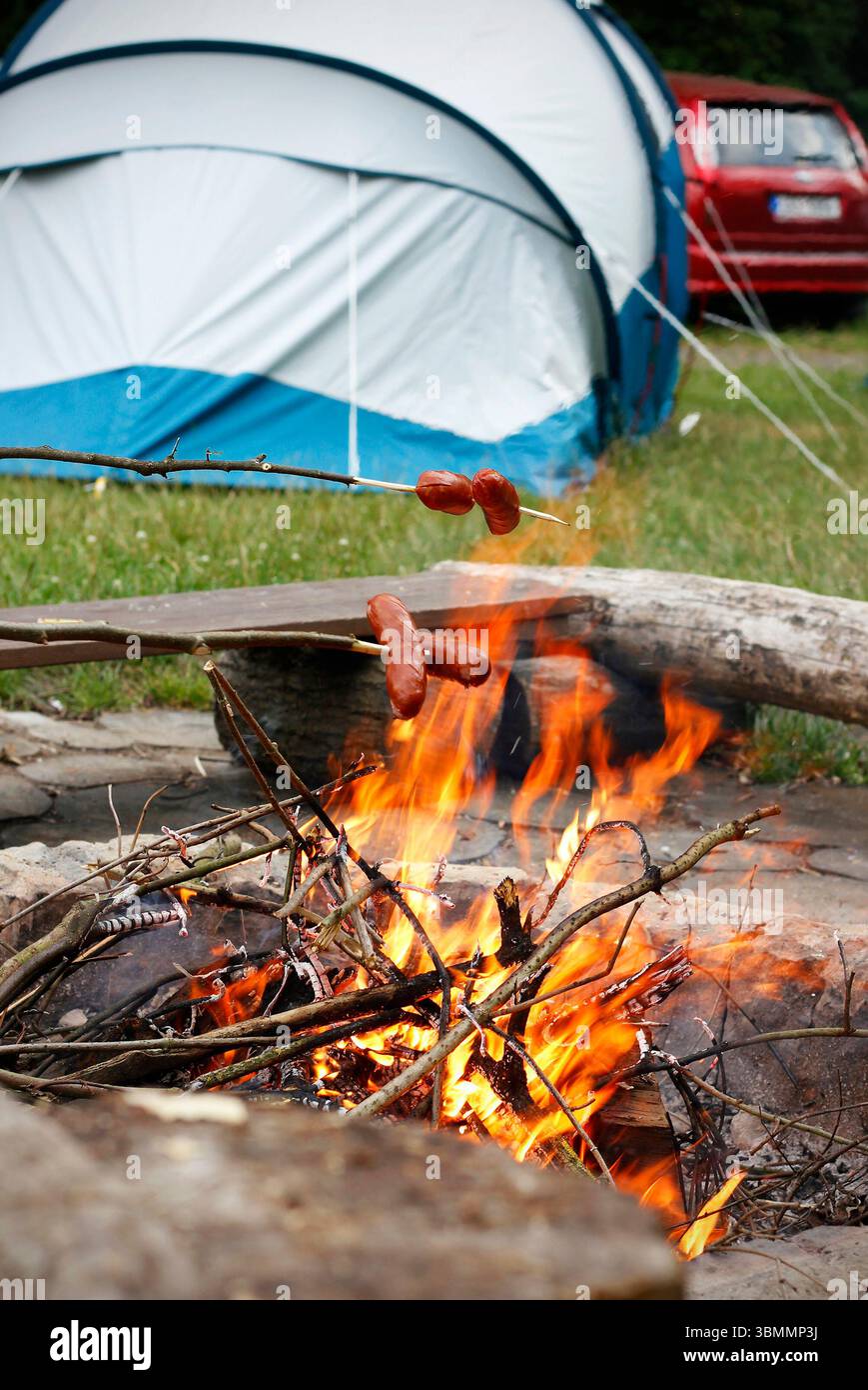 Sedmihorky, Repubblica Ceca - 07 20 2024: Salsicce alla griglia su una fiamma libera in un campeggio con tenda e auto sullo sfondo Foto Stock