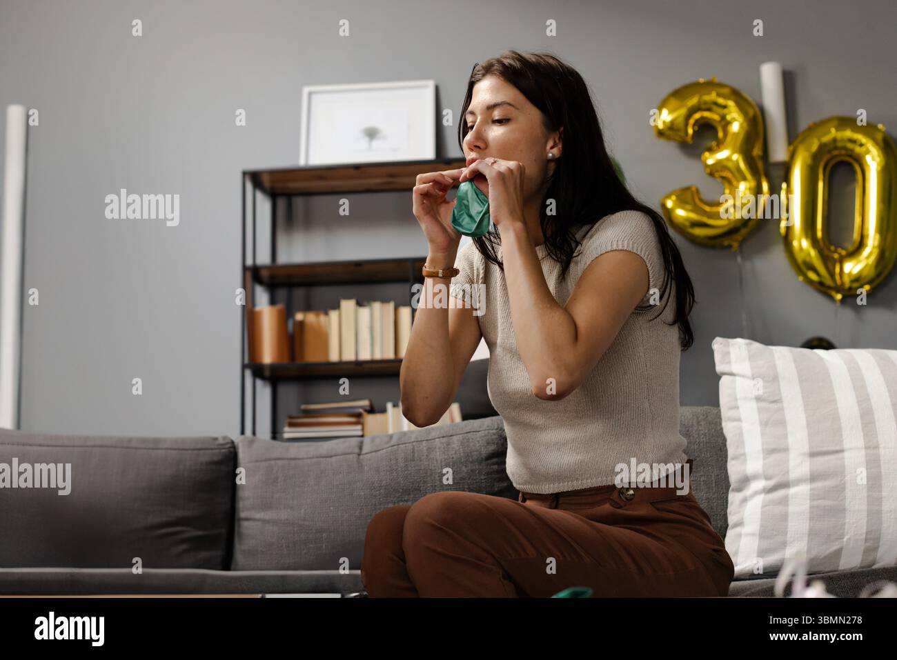 Donna seduta su un divano grigio a casa gonfiando palloncino verde vicino a palloncini dorati 30, spazio copia Foto Stock