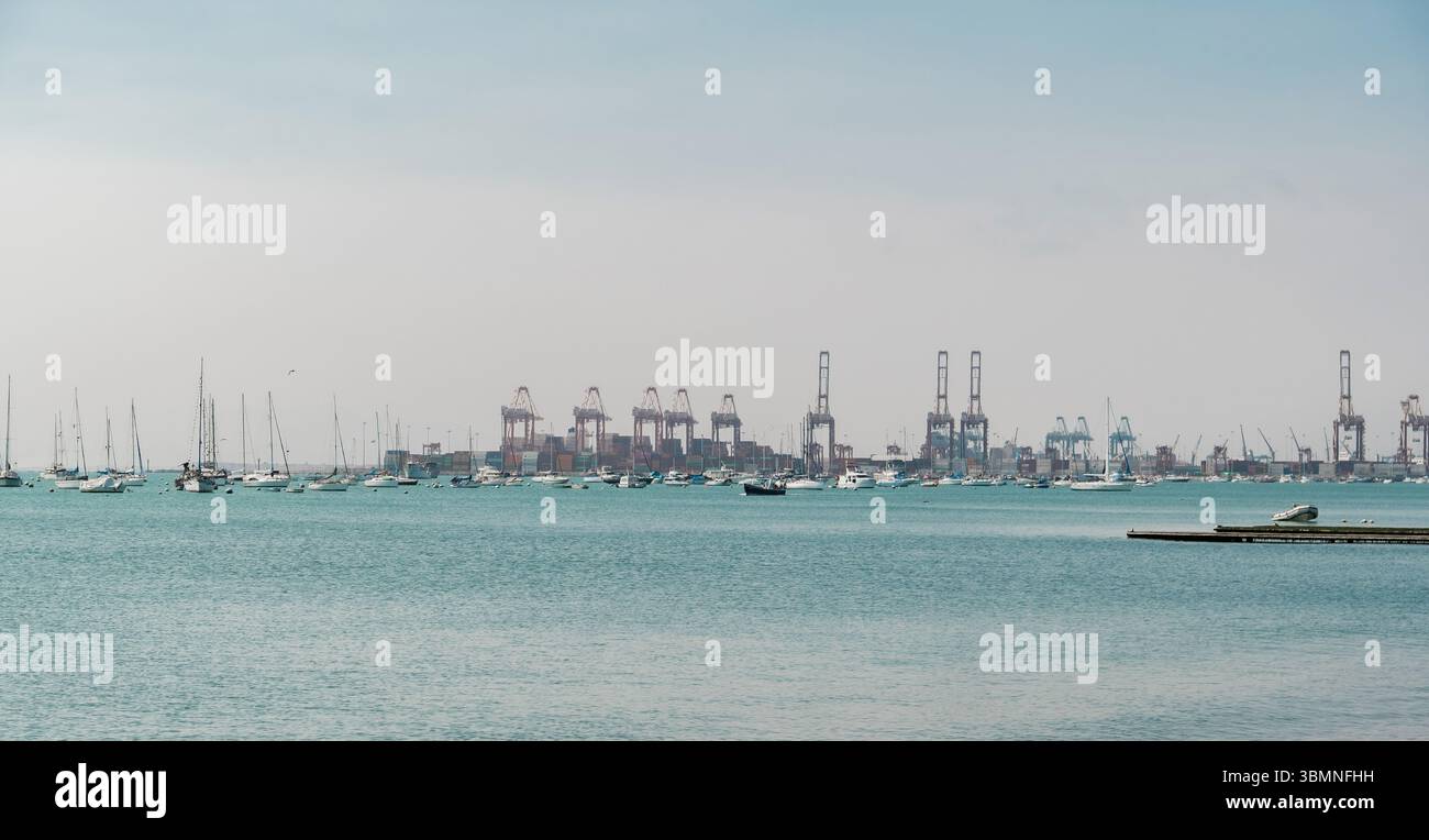Baia tranquilla con barche a vela e navi portacontainer che condividono l'acqua con grandi gru industriali che dominano lo skyline Foto Stock