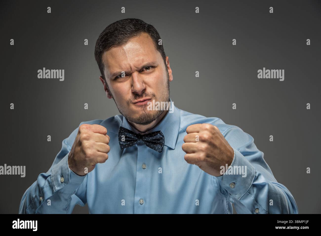 Ritratto di un giovane arrabbiato con camicia blu e cravatta a farfalla con pugni isolati su sfondo grigio studio. Emozione umana negativa, esp facciale Foto Stock