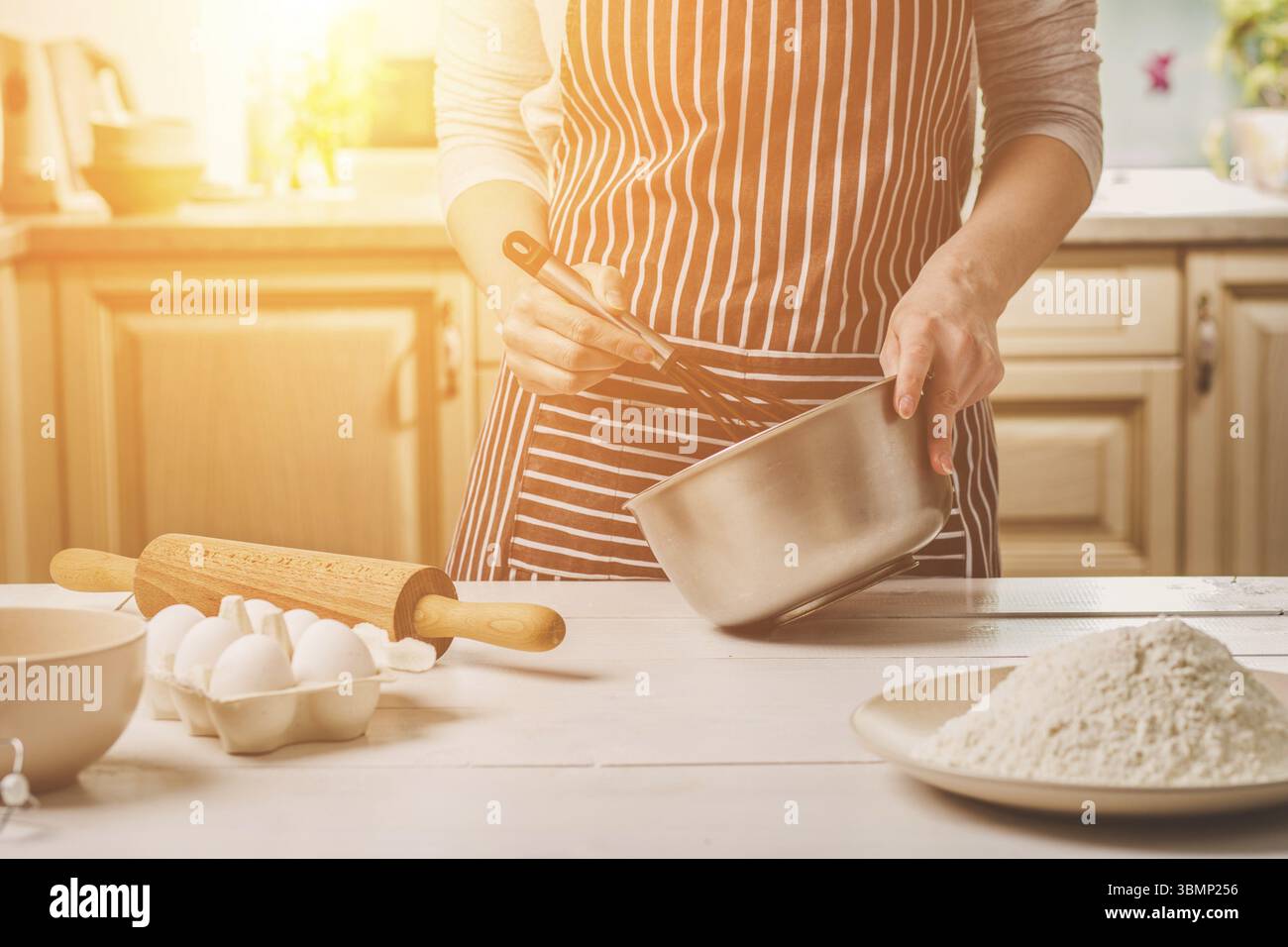 Giovane donna tenendo la coppa con impasto e mescolare con una frusta, primo piano. Una donna in un grembiule a strisce è la cucina in cucina Foto Stock