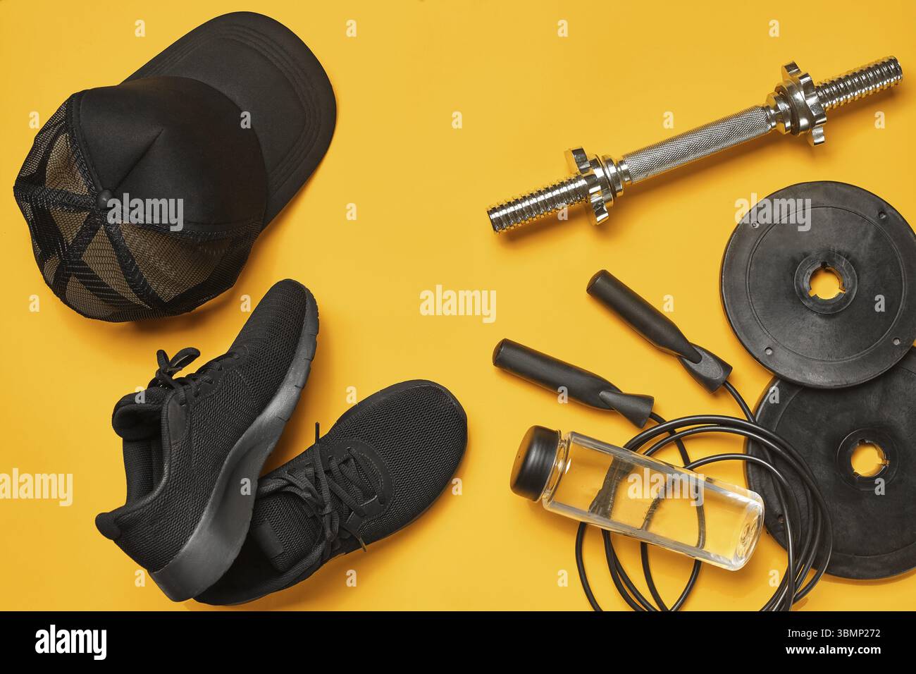 Primo piano di un accessorio nero da palestra, come scarpe sportive, bottiglia trasparente, manubri, berretto da baseball e corda per saltare su sfondo giallo. T Foto Stock