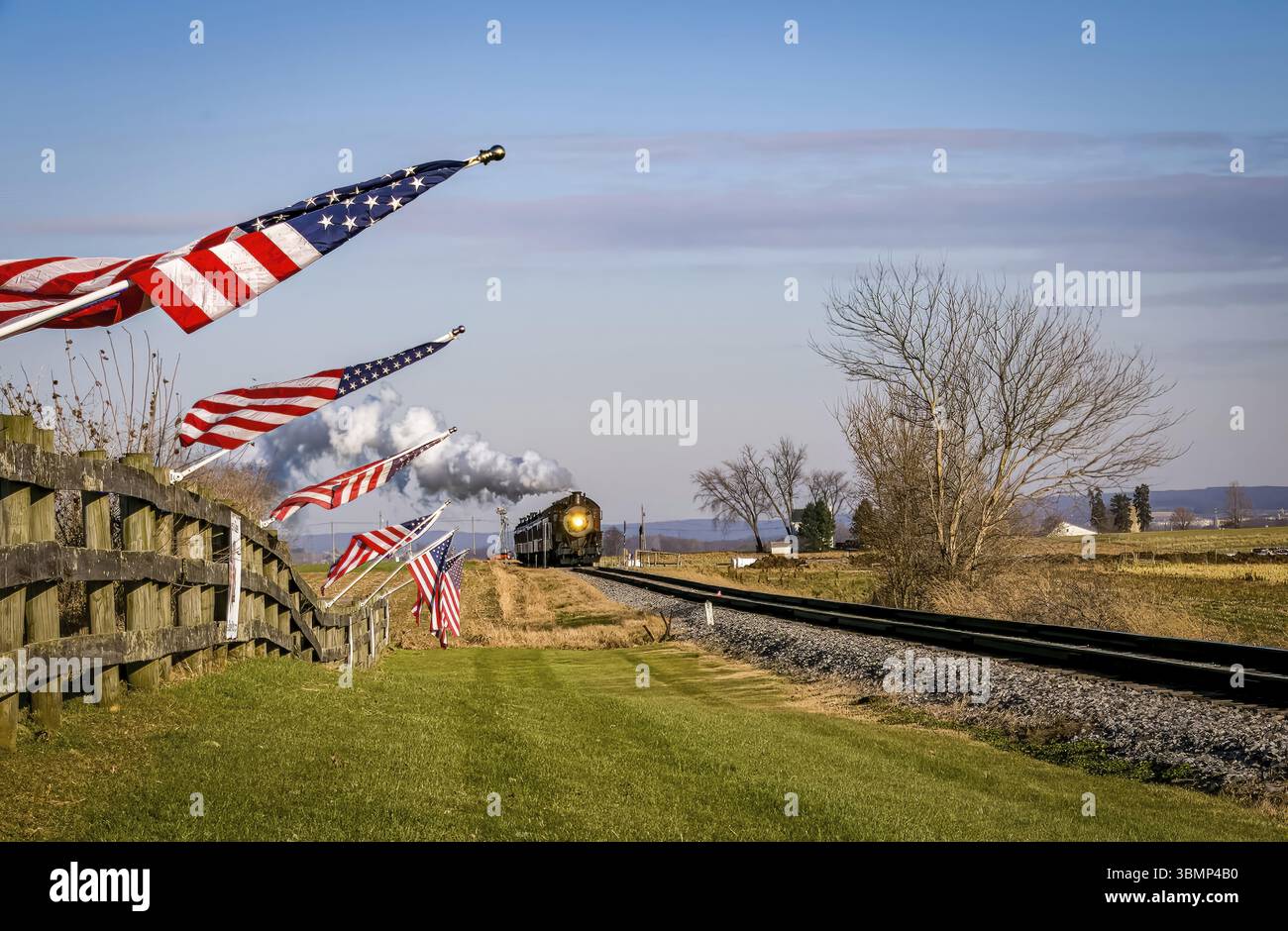 Una veduta di un treno passeggeri a vapore classico che si avvicina, con le bandiere americane attaccate ad una recinzione in un giorno estivo soleggiato Foto Stock