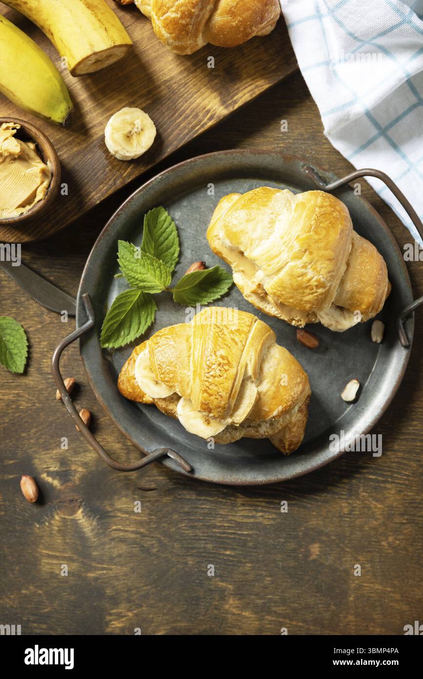 Concetto di dessert di cibo sano. Pasticceria francese. Gustosi croissant appena sfornati con burro di arachidi e banana su uno sfondo rustico. Vista dall'alto. Poliziotto Foto Stock
