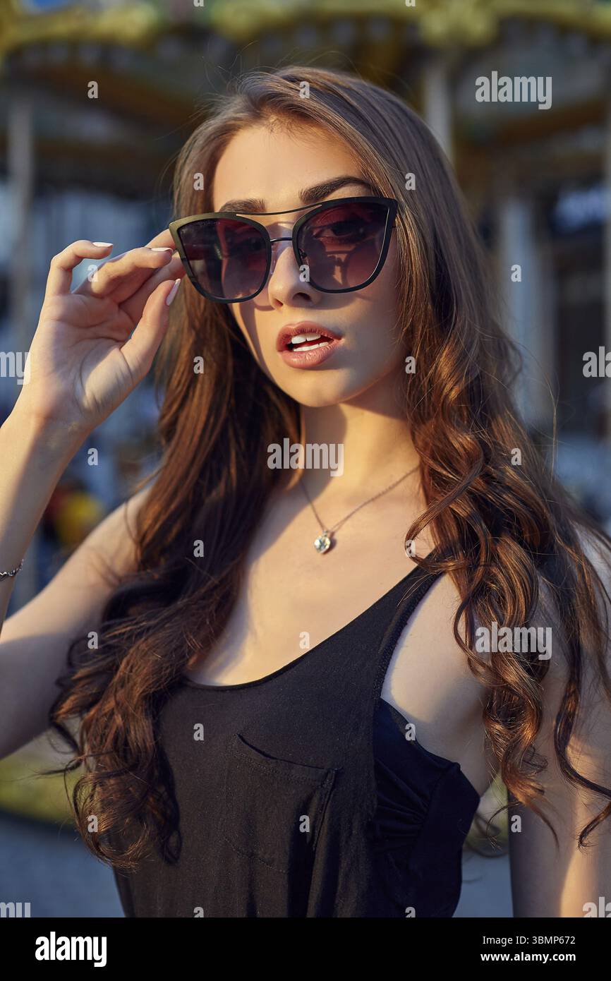 Ritratto di una bella donna bruna in abito da sera accanto a una giostra per bambini. Ritratto in stile moda di giovane bella donna elegante in blac Foto Stock