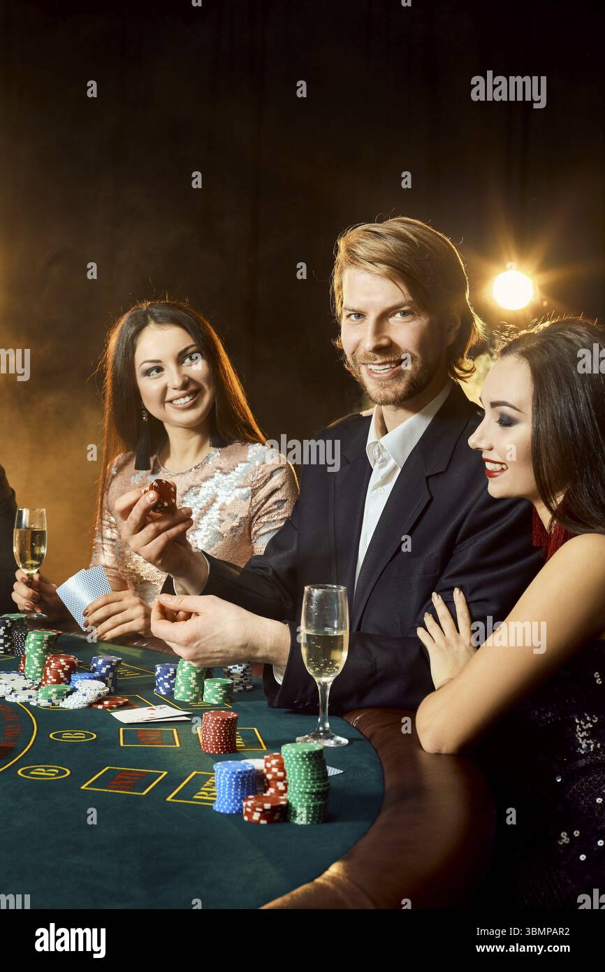 Un gruppo di giovani persone ricche è giocare a poker nei casinò. Uomo in business suit e due giovani donne in nero abiti. Il fumo. Casino. Poker Foto Stock