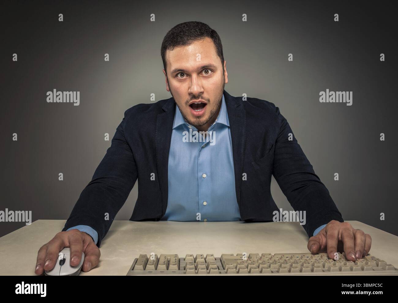Ritratti il giovane uomo con la camicia blu e la giacca che guarda la fotocamera con stupore, seduto a una scrivania vicino a un computer isolato su uno sfondo grigio di studio Foto Stock