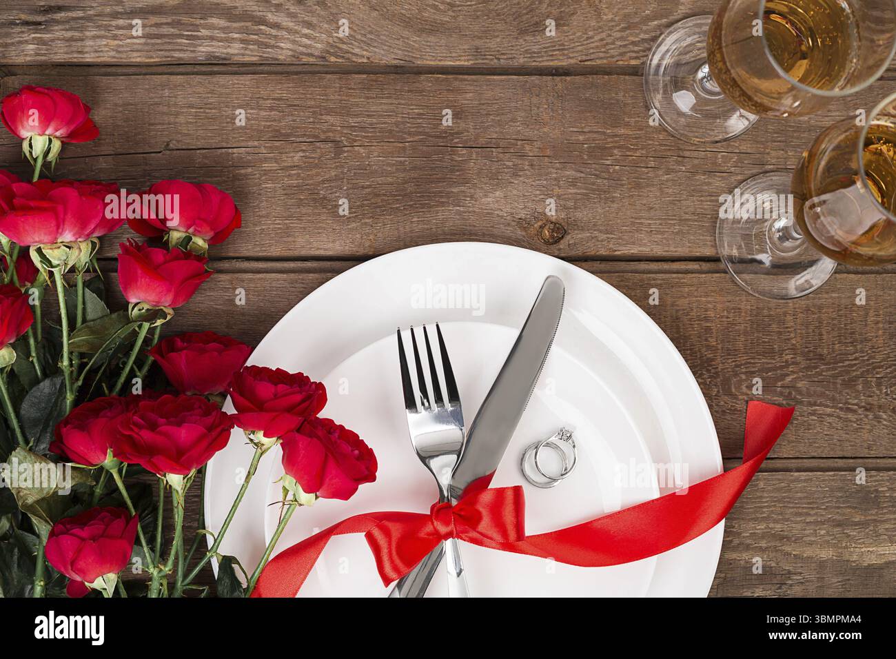 Cena di San Valentino messa in tavola con nastro rosso, rose, coltello e forchetta e anello su sfondo di quercia. Still Life Foto Stock