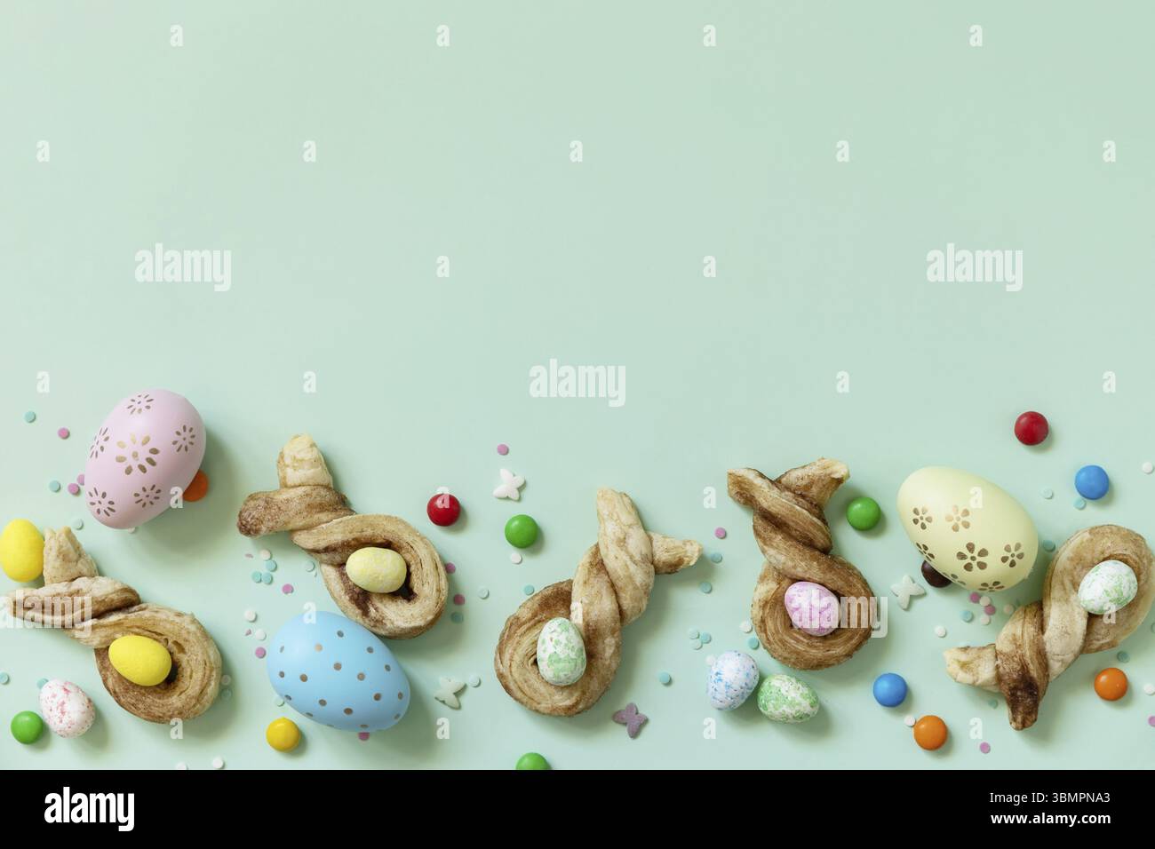 Colori uova di Pasqua con focaccine di Pasqua a forma di coniglio pasta sfoglia con cannella su fondo verde pastello. Buone feste di Pasqua. Vista dall'alto. Copia Foto Stock