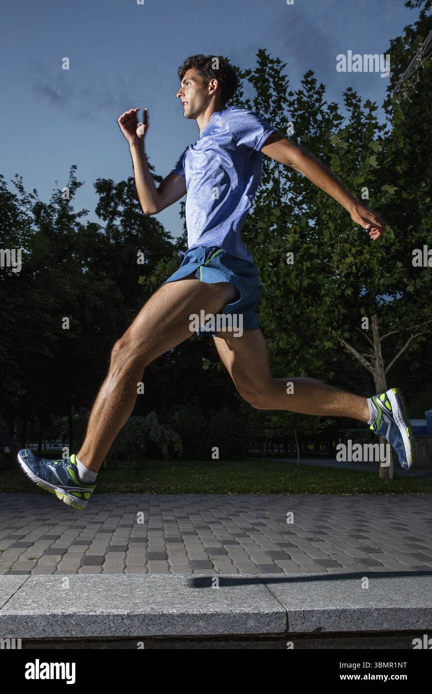 Uomo che corre nel parco della città al tramonto. Abbigliamento sportivo blu, pantaloncini e maglietta Foto Stock