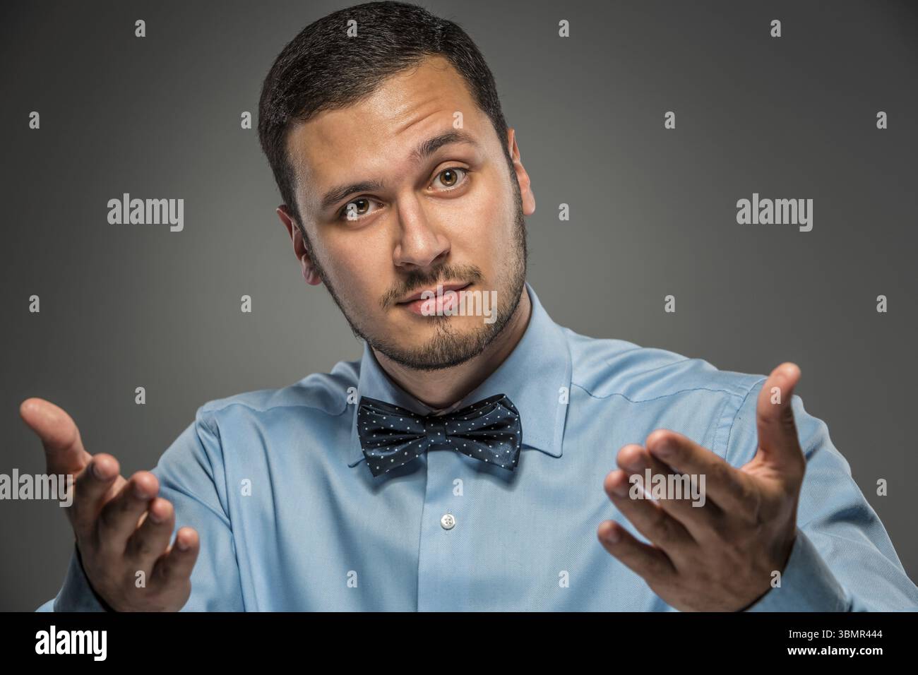 Ritratti il giovane uomo in camicia blu e cravatta a farfalla che guarda la fotocamera, mani alzate, isolato su sfondo grigio studio. Emozione umana, espr facciale Foto Stock