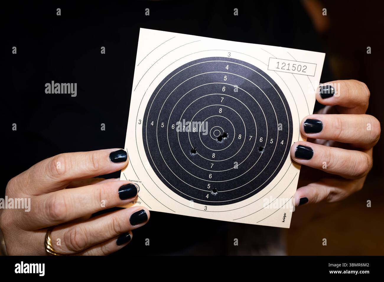 Landkreis Osnabrück, Deutschland 25. Giu 2025: Im Bild: Zwei Hände mit dunklem Nagellack halten eine klassische Papierscheibe, die nach einer Schussserie im 50-Meter-Kleinkaliber KK-Schießen verwendet wurde. Auf der Scheibe sind mehrere Trefferlöcher sichtbar, Die das Schießergebnis einer Athletin oder eines Athleten dokumentieren. Die Ringeinteilung der Scheibe, von außen nach innen nummeriert, Dient der Bewertung der Präzision. Die Seriennummer 121502 ist oben rechts auf der Scheibe vermerkt. Dieses Bild zeigt das unmittelbare Ergebnis des Präzisionsschießens und die greifbaren Resultate de Foto Stock