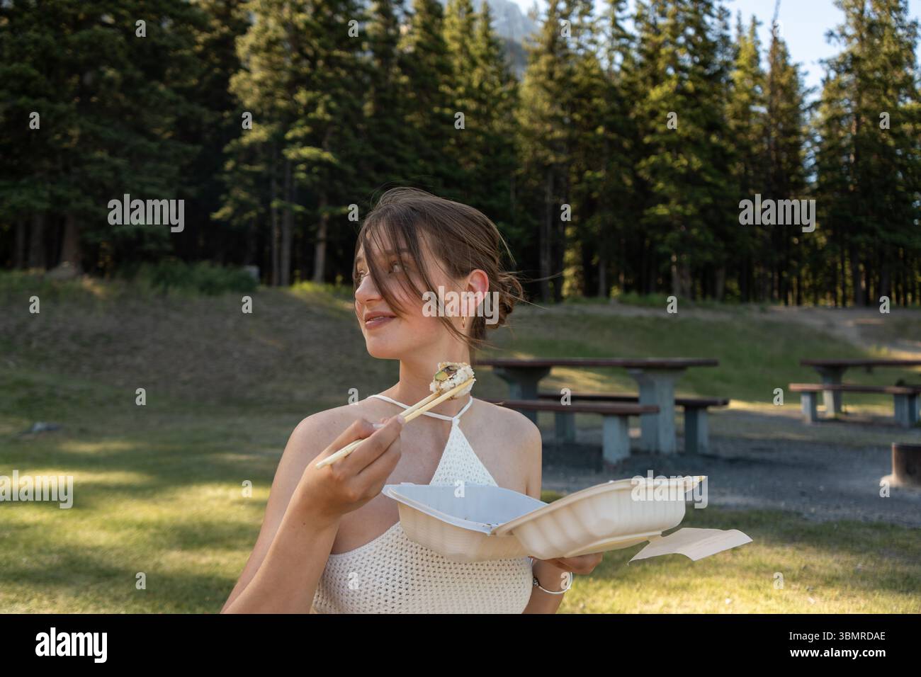 Donna sorridente che tiene il sushi con le bacchette nel soleggiato parco all'aperto Foto Stock