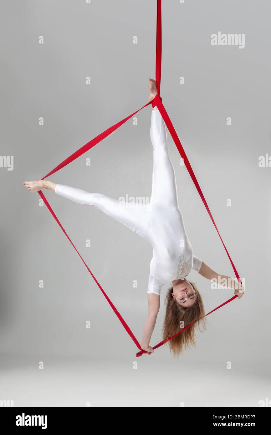 Esibizione ginnasta su tele rosse. Una bella ragazza con una tuta sportiva leggera sta facendo degli elementi acrobatici appesi a testa in giù su una corda in uno studio i Foto Stock