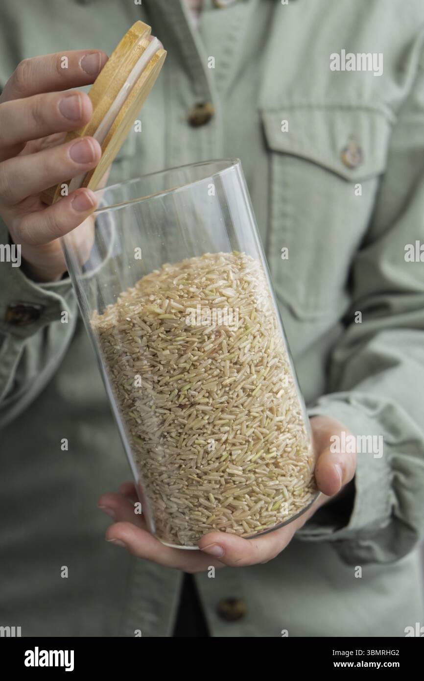 Donna irriconoscibile che tiene in mano un vaso di vetro ricaricabile con porridge di riso crudo. Ricarica formato prodotto ecologico. Riempimento del contenitore riutilizzabile Foto Stock