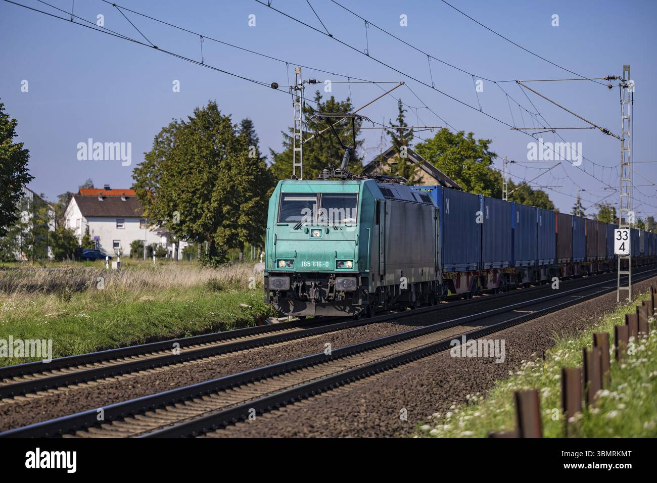 Un treno merci turchese su una linea ferroviaria elettrica che attraversa un'area rurale vicino a un insediamento per il trasporto pubblico e il traffico regionale di passeggeri Foto Stock