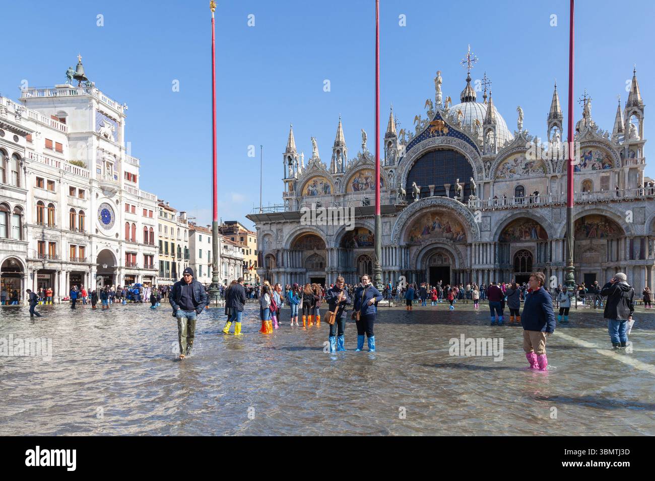 I turisti e la gente del posto camminano attraverso acqua alta marea, Piazza San Marco, Venezia, Italia con la Basilica di San Marco e la Torre dell'Orologio, cambiamenti climatici Foto Stock