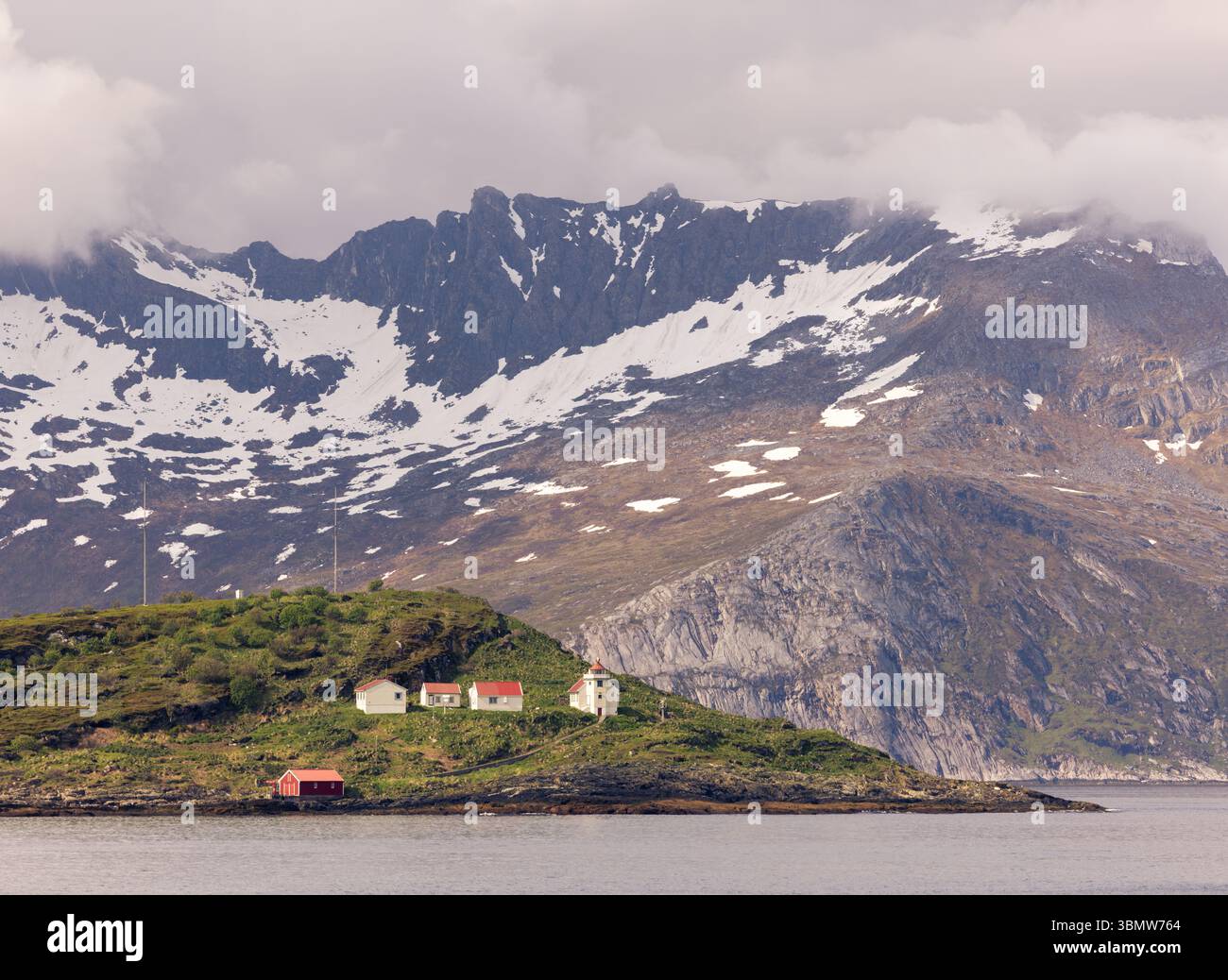 Faro e piccole case su un'isola remota nella Norvegia artica, con spettacolari montagne spolverate di neve alle spalle e condizioni di disgelo primaverile. Foto Stock