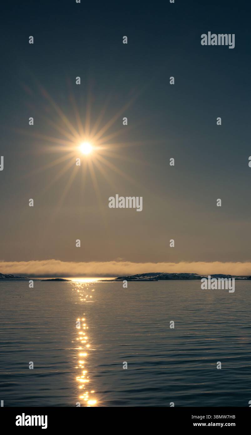 Sole di mezzanotte sopra il calmo mare Artico vicino a Sisimiut, in Groenlandia. Il sole luminoso si riflette sull'acqua con nebbia bassa e isole lontane. Foto Stock