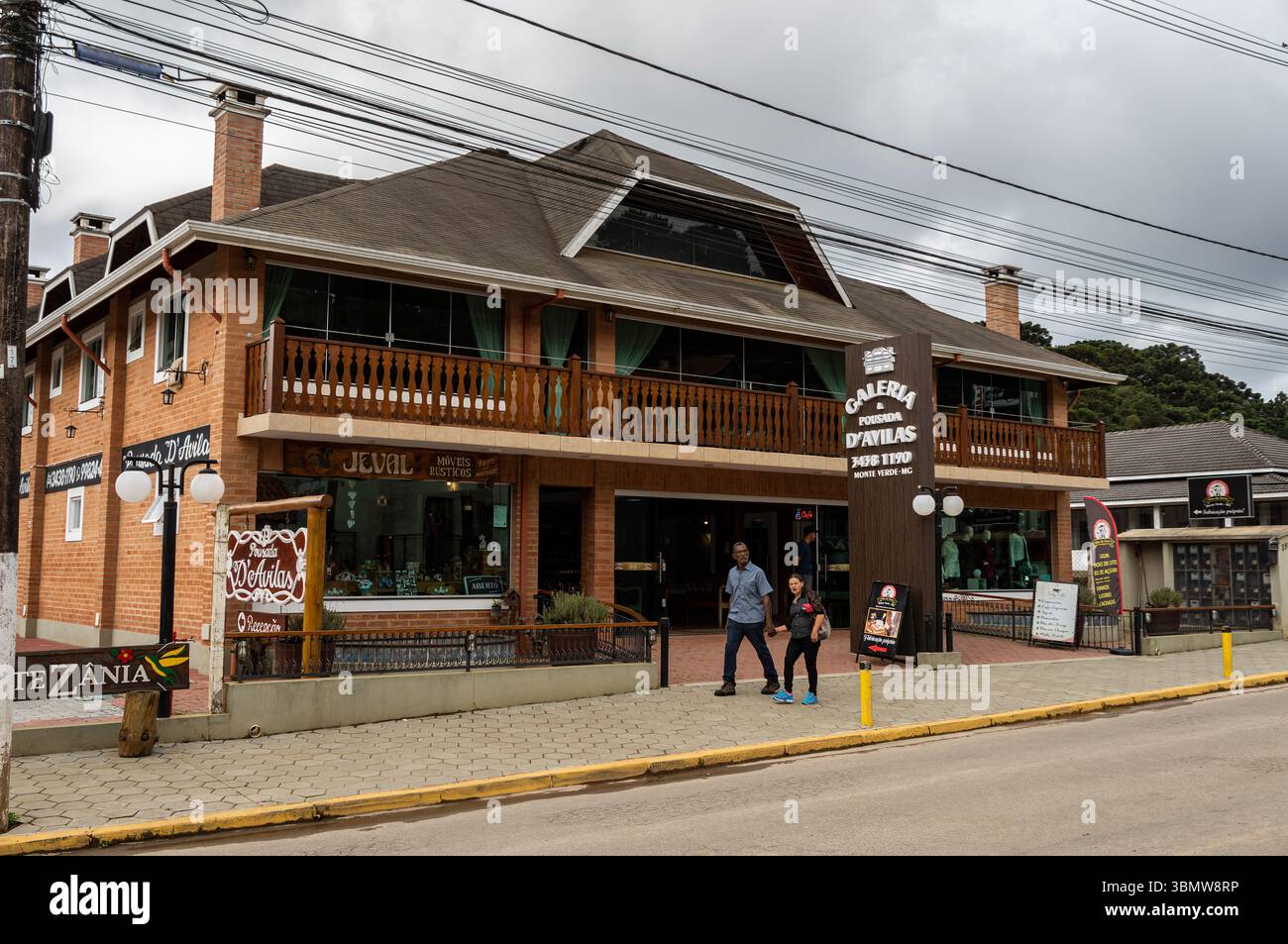 Facciata in mattoni della Galeria Pousada D'Avilas con negozi e pedoni che camminano sul marciapiede del viale principale del Monte Verde sotto il cielo nuvoloso. Foto Stock