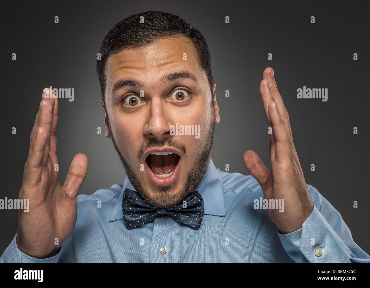 Ritratti il giovane uomo con la camicia blu e la cravatta a farfalla guardando la fotocamera con stupore, isolato su sfondo grigio studio. Emozione umana, esp facciale Foto Stock