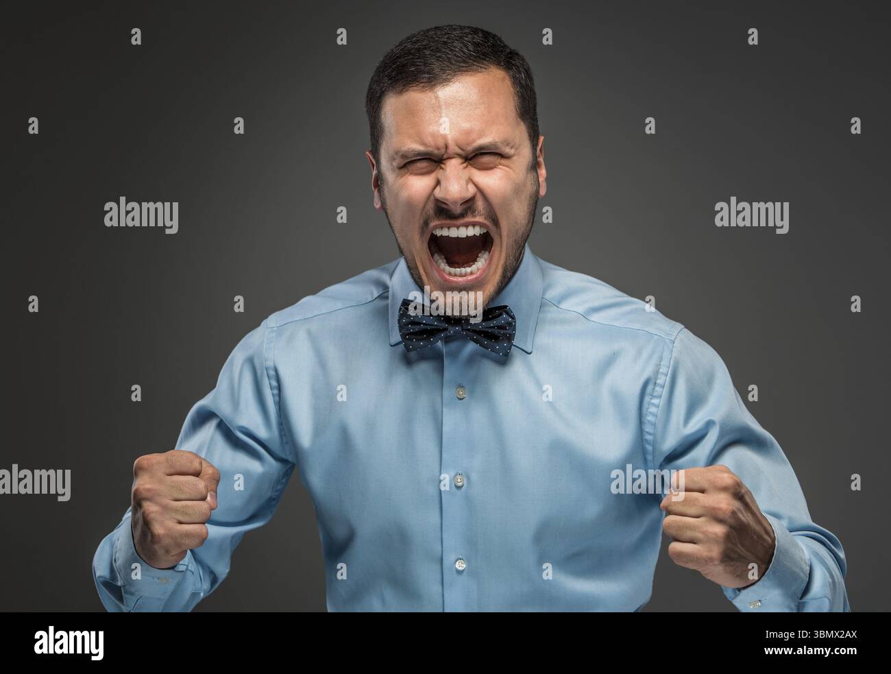 Ritratto di un giovane arrabbiato con camicia blu e cravatta a farfalla con pugni urlanti isolato su sfondo grigio studio. Emozione umana negativa, fa Foto Stock