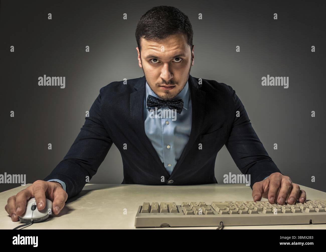 Ritratti il giovane uomo con la camicia blu e la giacca che guarda la fotocamera, seduto a una scrivania vicino a un computer isolato su uno sfondo grigio dello studio. Emozione umana Foto Stock