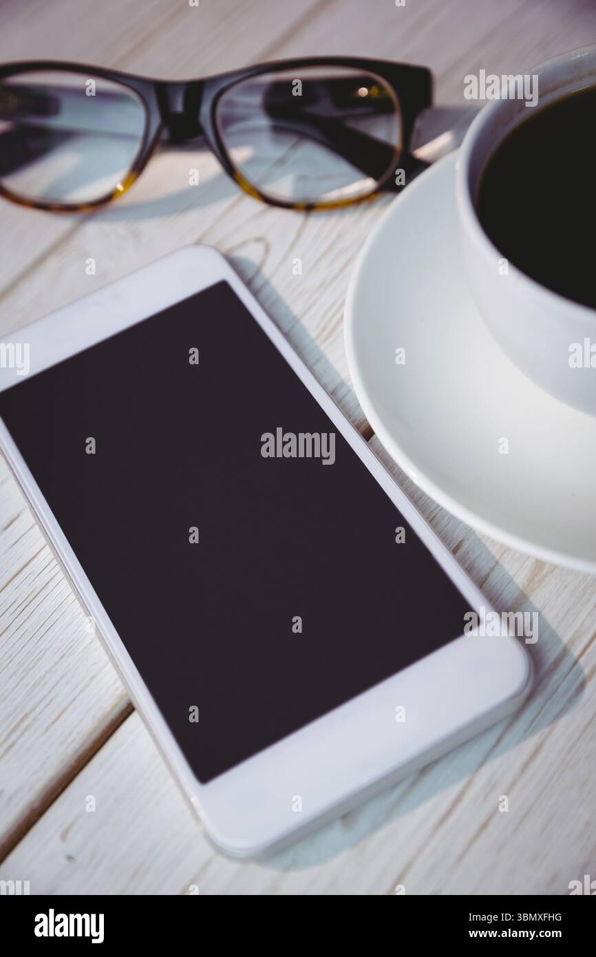 Piano in legno chiaro con smartphone bianco, occhiali con cornice nera e tazza da caffè Foto Stock