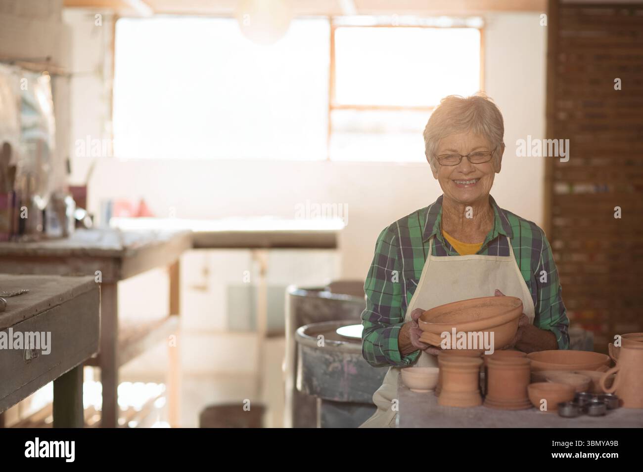 Donna anziana che indossa un grembiule che tiene una ciotola di argilla alla ruota di ceramica sorridendo in studio, spazio copia Foto Stock