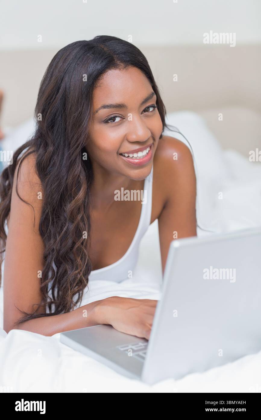 Donna afroamericana che indossa canotta bianca adagiata su biancheria bianca utilizzando un computer portatile argento in camera da letto Foto Stock