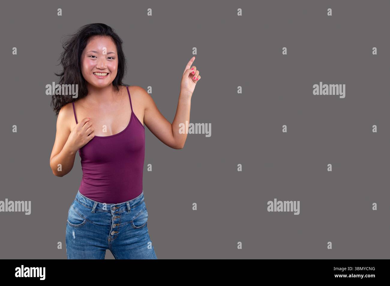 Giovane donna allegra in un outfit casual che punta verso l'alto con un sorriso, isolata su sfondo grigio per pubblicità o spazio messaggi. Foto Stock