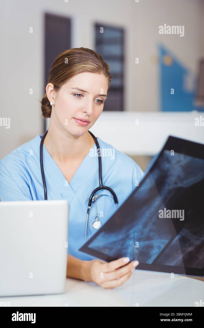 Medico donna che indossa scrub e stetoscopio, che tiene la pellicola a raggi X, controlla il computer portatile alla scrivania della clinica Foto Stock