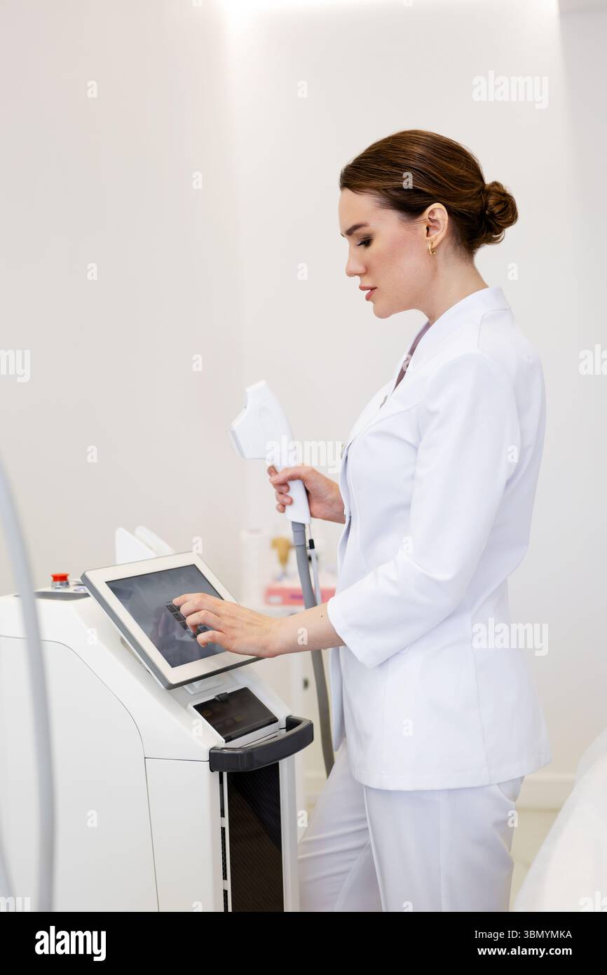 Donna cosmetologa in giacca bianca dispositivo laser di regolazione prima di procedura non chirurgica di ringiovanimento della pelle in clinica di bellezza, attrezzature moderne per aesthe Foto Stock