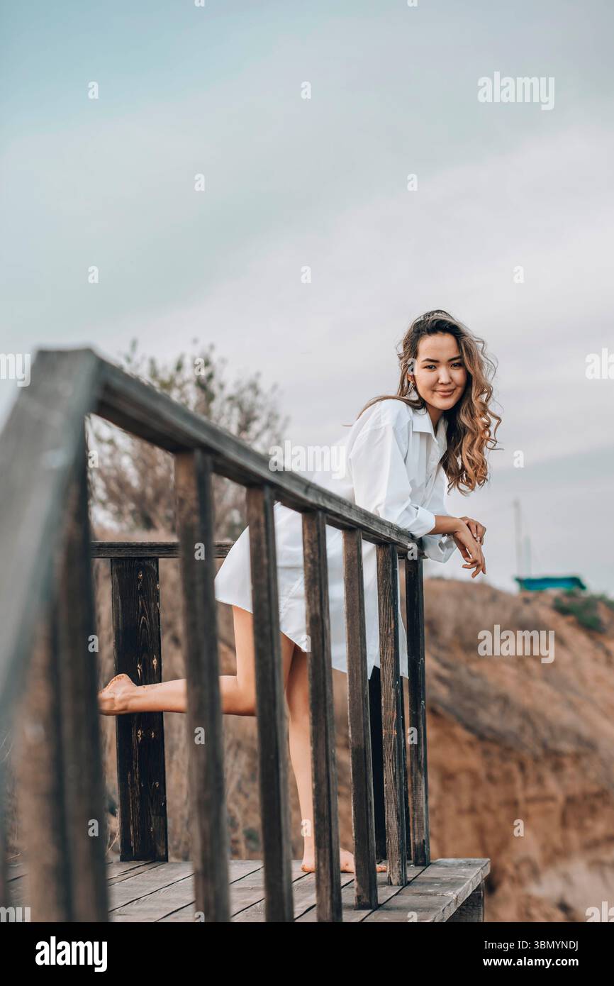 Donna asiatica in camicia bianca in piedi nudi su una piattaforma di legno con un piede sollevato, paesaggio roccioso naturale, vista sul retro, solitudine, personale Foto Stock